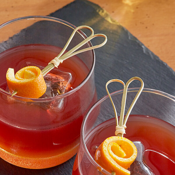 Two glasses of a cocktail garnished with orange spirals on a table with bamboo heart skewers.