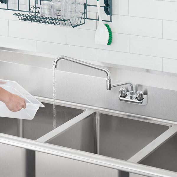 A person using a Regency wall mount faucet to pour water into a stainless steel sink.