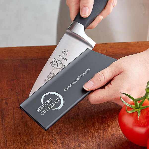 A hand holding a Mercer Culinary knife with a polypropylene blade guard on the counter cutting a tomato.