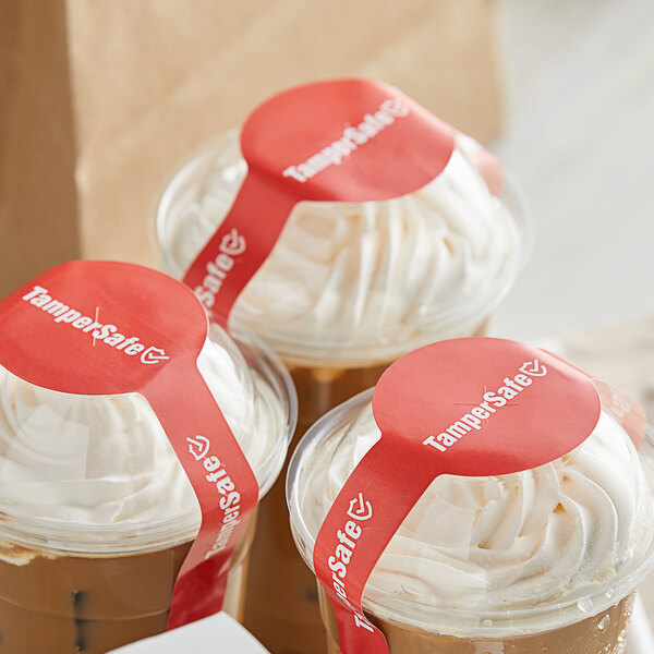 A group of plastic cups with TamperSafe red dome lids and drink labels filled with coffee and whipped cream.