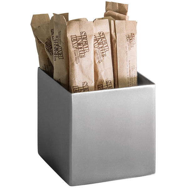 A silver brushed stainless steel square sugar caddy holding several brown bags of sugar.