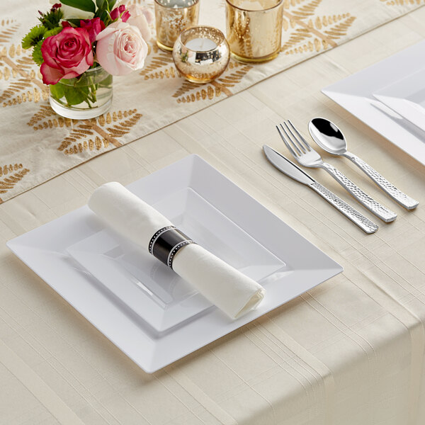 A white table setting with Visions white plastic dinnerware and Classic hammered flatware on a table with a rose in a glass vase.