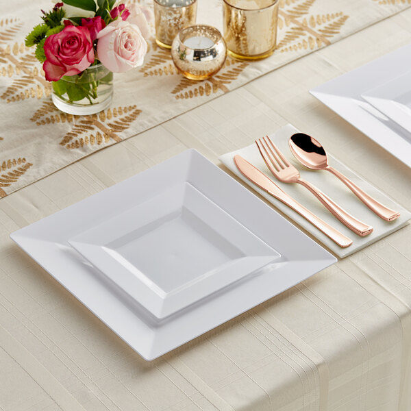 A table set with white square plastic plates, silver and gold plastic utensils.