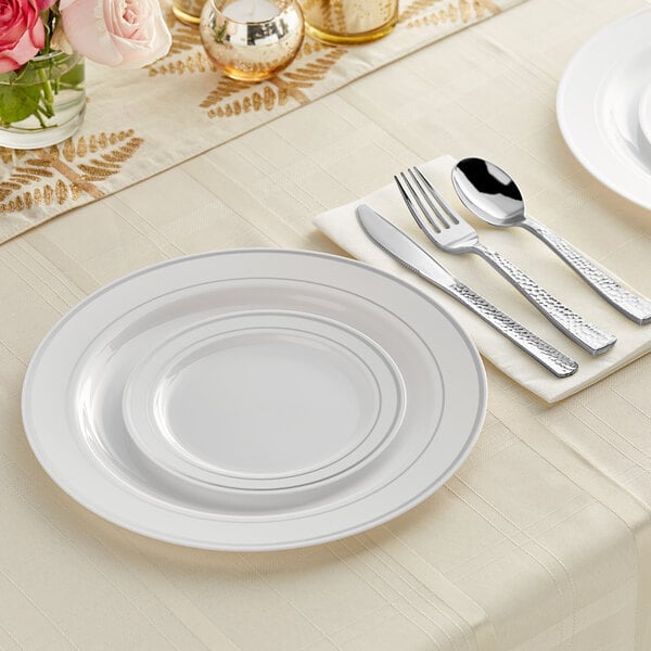 A white table setting with Visions banded plastic dinnerware and hammered flatware on a white table with roses.