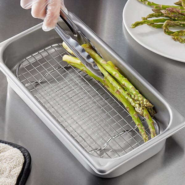 A person using tongs to serve asparagus in a stainless steel tray with a Vigor footed pan grate.