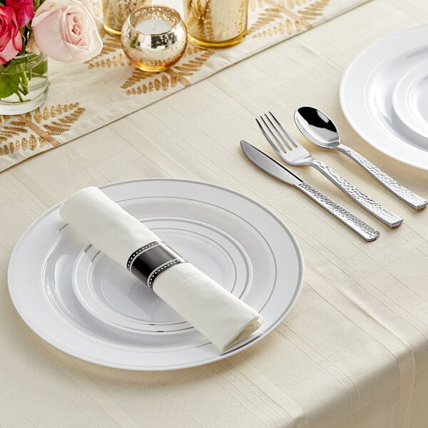 A white table setting with Visions silver banded plastic dinnerware and rolled hammered flatware on a white plate with a napkin.