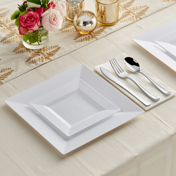 A white Visions square plastic plate with silver Classic flatware on a white surface with flowers.