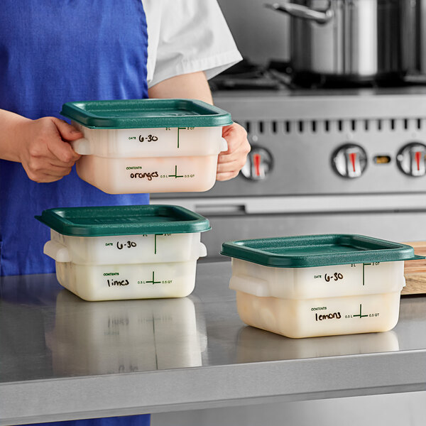 A woman in a blue apron holding Carlisle square plastic food storage containers with green lids.