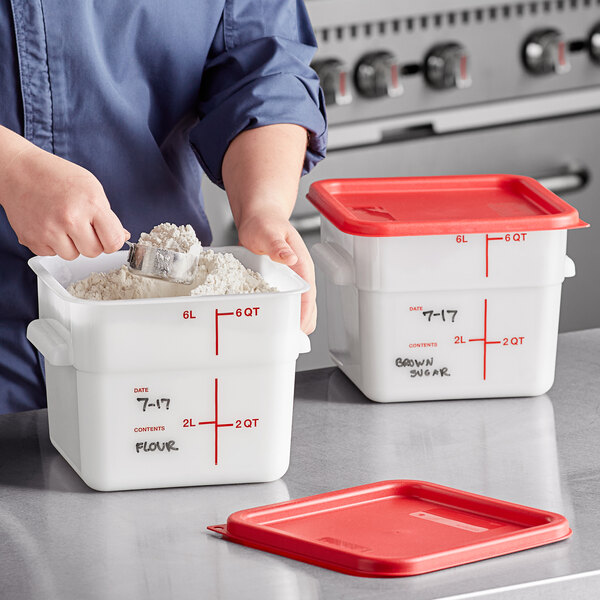 A person using a measuring cup to pour flour into a Carlisle white plastic food storage container with a red lid.