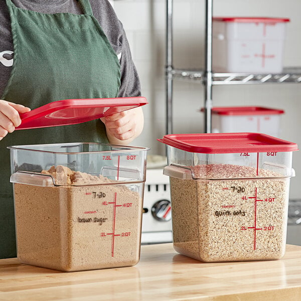 A person in an apron holding a red lid over a couple of Carlisle clear food storage containers.
