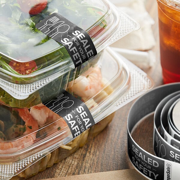 A roll of black TamperSafe labels with white writing on a stack of plastic containers filled with food.