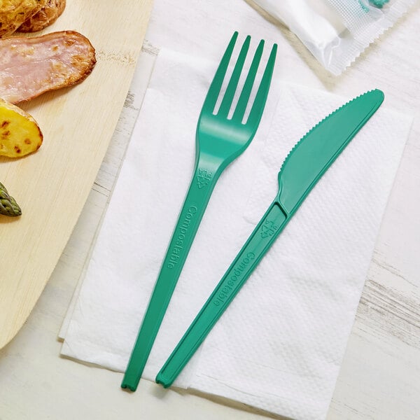 A green EcoChoice CPLA fork and knife on a napkin next to a plate of food.