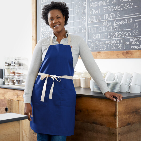 A woman wearing a Choice royal blue apron with natural webbing accents standing next to a blackboard.
