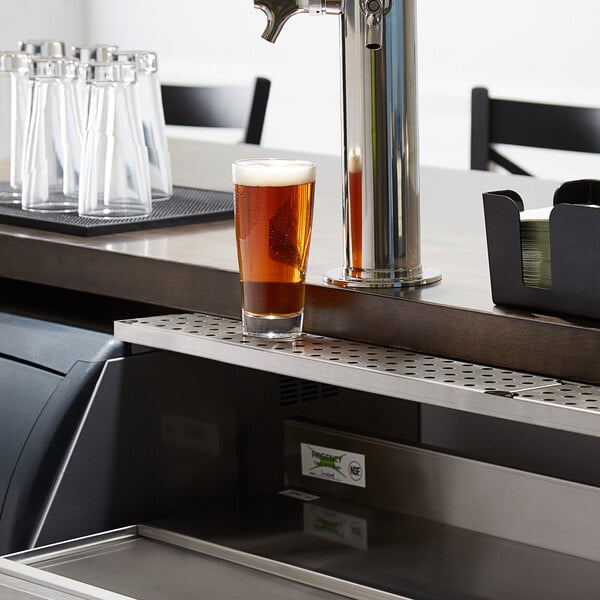 A glass of beer on a stainless steel underbar mount beer drip tray.