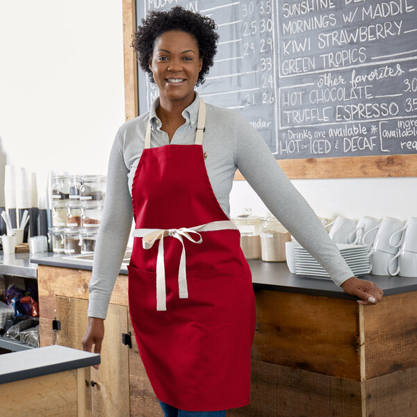 A smiling woman wearing a red Choice bib apron with natural webbing accents standing at a counter.