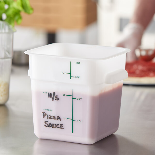 A hand using a measuring cup to pour pizza sauce into a Carlisle white square food storage container.