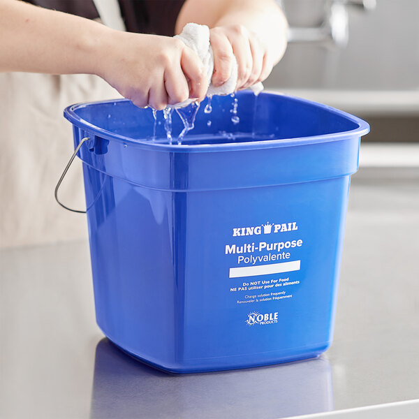 A person washing a Noble Products blue pail with a sponge.