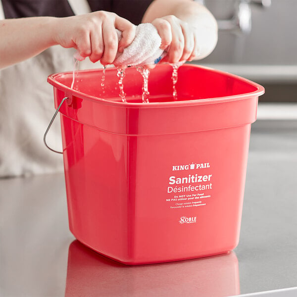 A woman washing a red Noble Products sanitizing pail on a counter in a school kitchen.