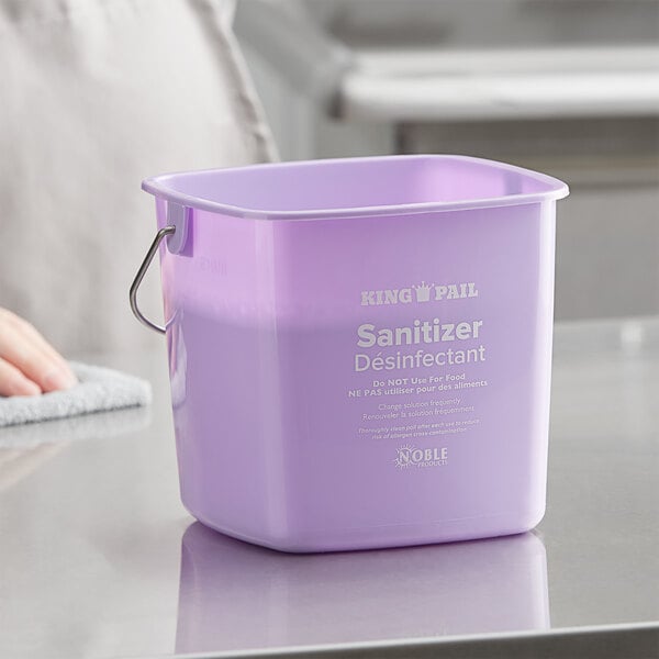 A person using a Noble Products purple sanitizing pail on a school kitchen counter.