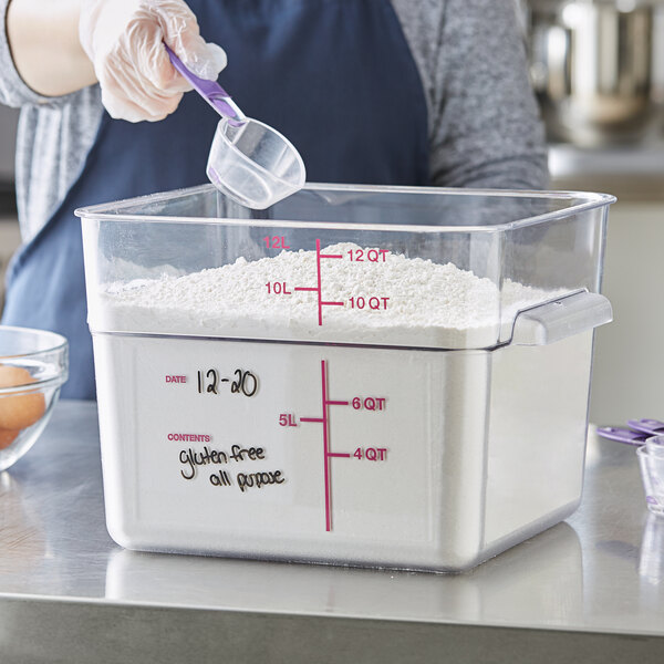 A person pouring flour from a measuring cup into a Carlisle clear polycarbonate food storage container.