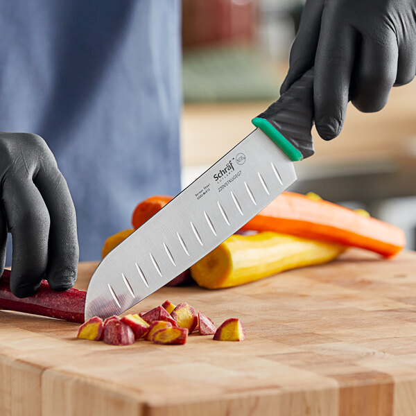 A person in black gloves using a Schraf Santoku knife with a green handle to cut a yellow vegetable.