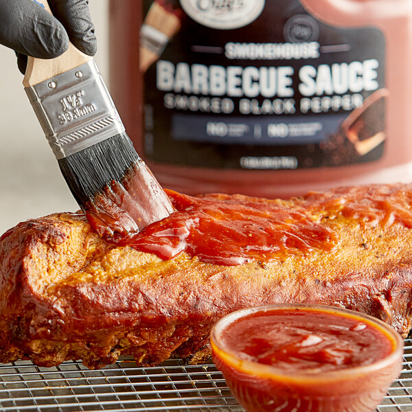 A hand using a brush to paint Sauce Craft Smoked Black Pepper BBQ Sauce on a rack of barbecue.