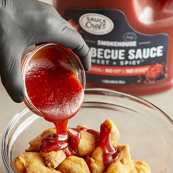 A gloved hand pours Sauce Craft Sweet and Spicy BBQ Sauce into a bowl of food.