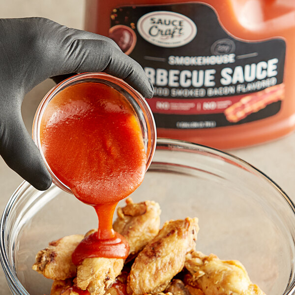 A person pouring Sauce Craft Applewood Smoked Bacon BBQ Sauce into a bowl of chicken wings.