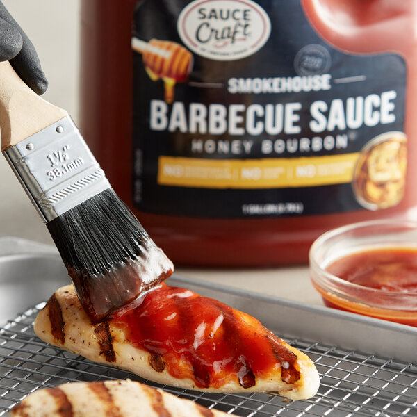 A person using a brush to paint Sauce Craft Honey Bourbon BBQ Sauce on a piece of chicken.