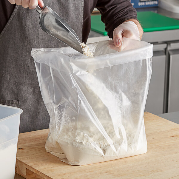 A hand using a metal scoop to put flour into a clear gusseted poly bag.