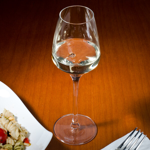 A Stolzle dessert wine glass filled with wine next to a plate of pasta and a fork.