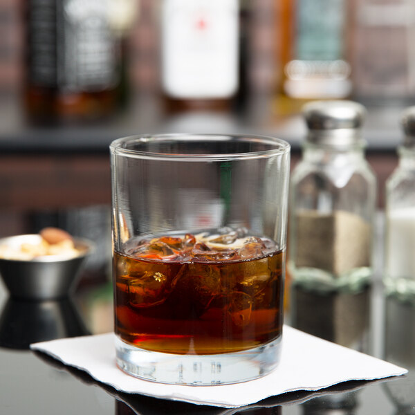 An Anchor Hocking Concord double old fashioned glass with brown liquid and ice on a napkin.