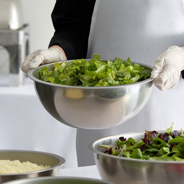 A person in a white apron holding a Vollrath stainless steel mixing bowl filled with green and red salad
