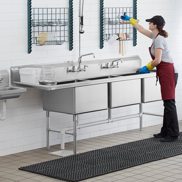 A woman wearing gloves cleaning a Regency stainless steel commercial sink.
