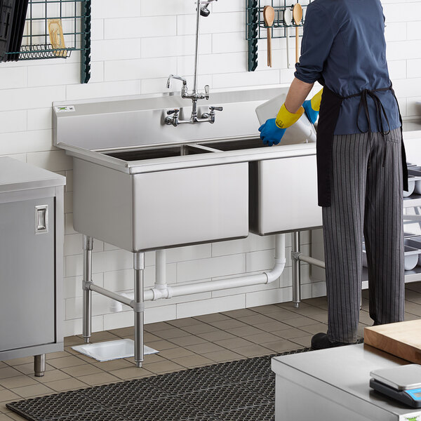 A person in a blue apron washing a Regency stainless steel two compartment sink.