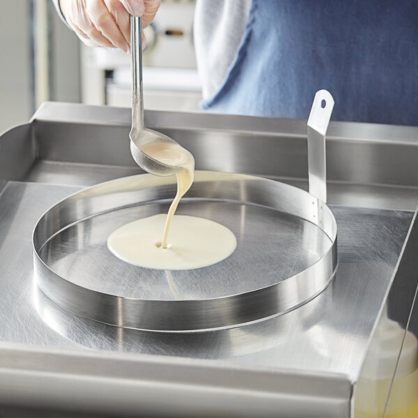 A hand using a spoon to pour liquid into a stainless steel egg ring in a pan.
