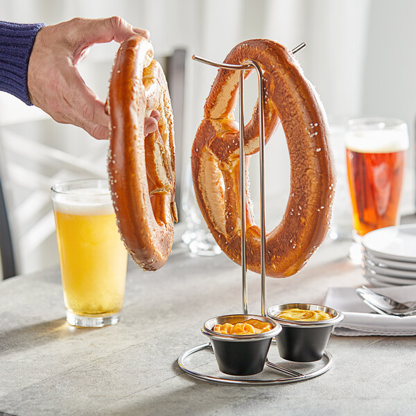 A hand holding a large pretzel over a Tablecraft stainless steel holder with dips.