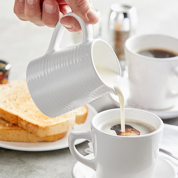 A person pouring milk into a white cup of coffee using a Tablecraft white melamine creamer.