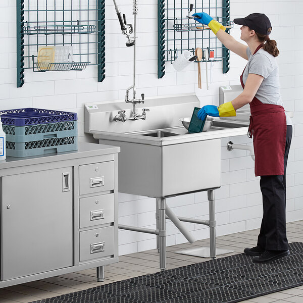 A woman wearing yellow gloves cleaning a Regency stainless steel commercial sink with a right drainboard.