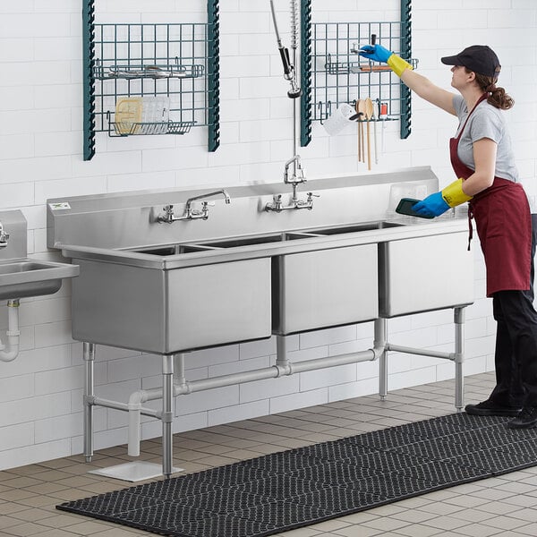 A woman wearing gloves cleaning a Regency stainless steel three compartment sink in a professional kitchen.