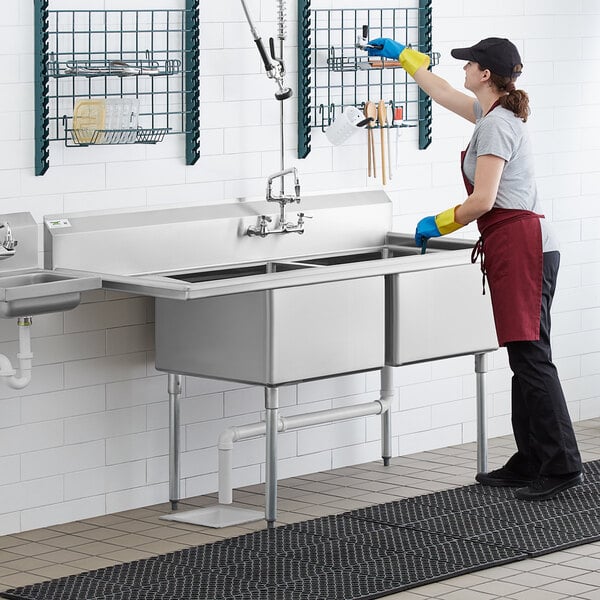 A woman in a black apron and gloves washing a Regency stainless steel two compartment sink.
