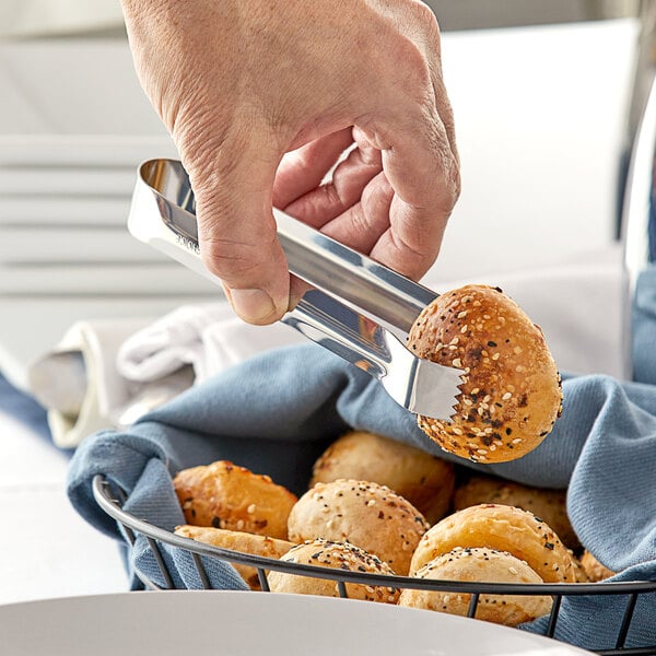 A person using Choice stainless steel pom tongs to pick up a bun from a basket.