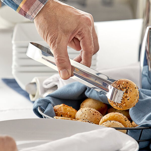 A hand holding Choice stainless steel tongs over a plate of bagels.