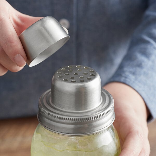 A person pouring lemonade from a Mason jar with a metal lid into a shaker.