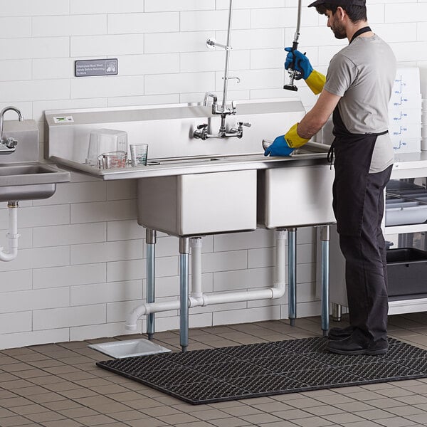 A person wearing gloves standing next to a Regency stainless steel two compartment sink with a drainboard.