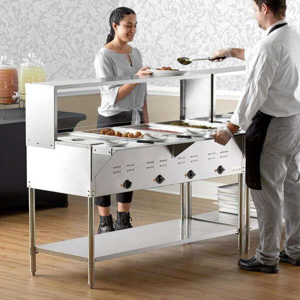 A man and woman cooking food on an Avantco electric steam table with sneeze guard.