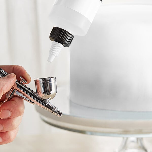 A person using a silver Chefmaster airbrush to spray a cake.