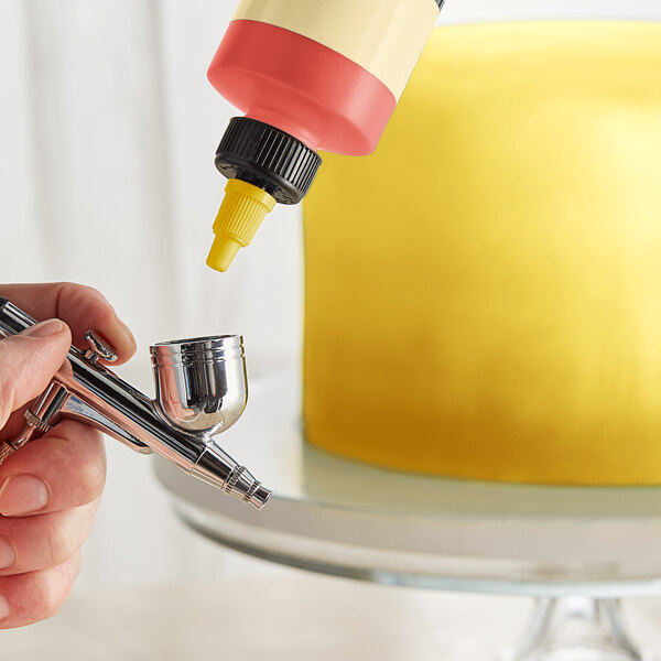 A hand using a silver airbrush to spray yellow color over a cake.