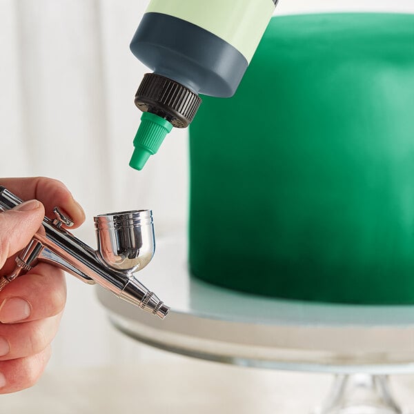 A person using a silver airbrush to paint a cake green with Chefmaster Neon Brite Green food coloring.