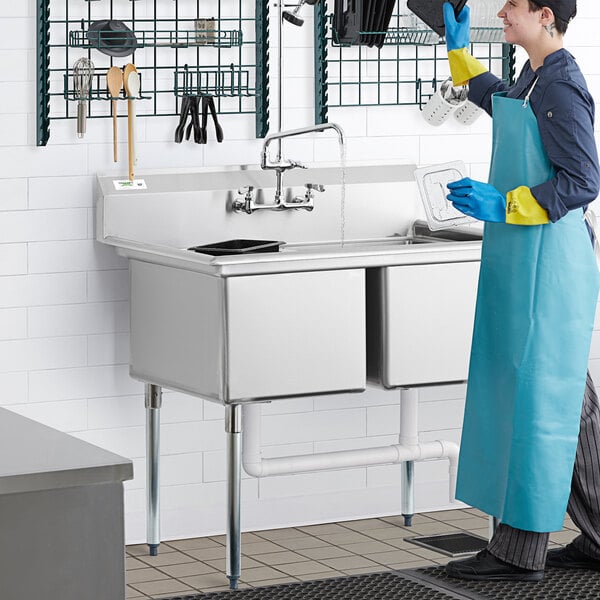 A woman in a blue apron washing dishes in a Regency two compartment sink.